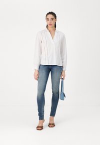 Hvit bluse med V-hals, subtil vertikal søm, lange ermer. Kombinert med blå skinny jeans og brune sandaler. Holder en liten blå veske.