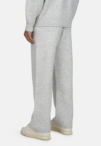 Kobieta ubrana w luźne jasnoszare dzianinowe spodnie i pasujący sweter, stoi w beżowych sneakersach widocznych na białym tle.