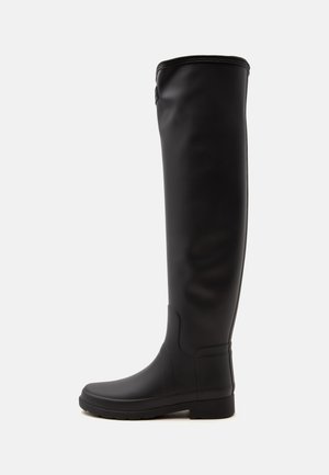 Bota alta negra hasta la rodilla con suela plana, acabado liso y punta redondeada, mostrada de lado sobre un fondo liso.