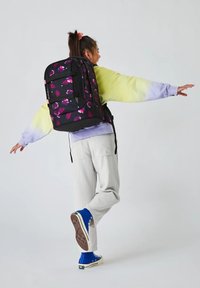 Sac à dos noir avec un motif floral rose, bretelles rembourrées et plusieurs compartiments. Porté sur le dos d'une personne, associé à des vêtements légers.