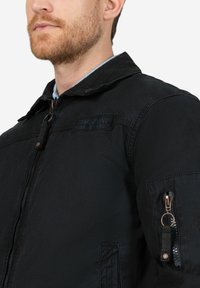 Schwarze Zip-Up-Jacke mit Kragen und vorderen Taschen. Sie verfügt über einen strukturierten Stoff, eine seitliche Reißverschlusstasche und ein Etikett auf der Brust.