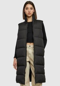 Veste longue noire en duvet avec un col haut, un design matelassé, une fermeture éclair à l'avant et des fentes latérales jusqu'en bas, portée sur un haut noir et un pantalon beige.