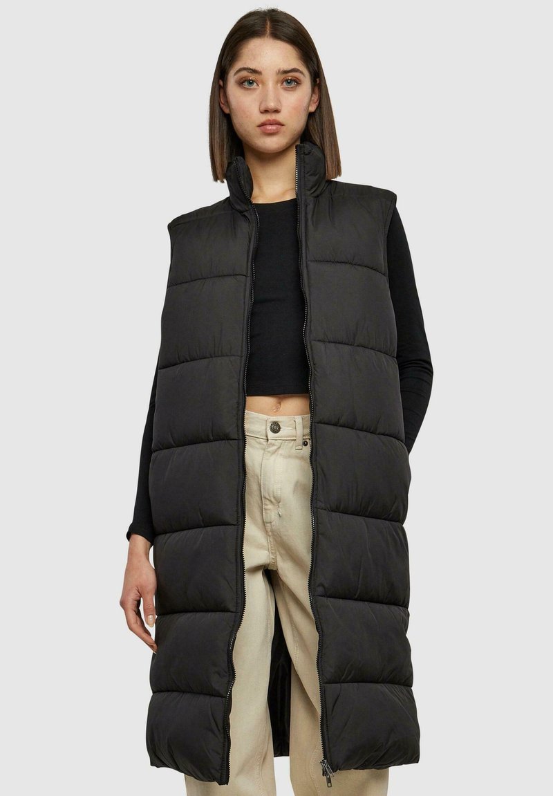 Veste longue noire en duvet avec un col haut, un design matelassé, une fermeture éclair à l'avant et des fentes latérales jusqu'en bas, portée sur un haut noir et un pantalon beige.