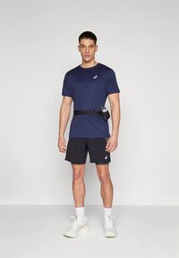 Marineblauw sport-T-shirt met een klein logo, gecombineerd met zwarte shorts. Een zwarte riem met een waterfleshouder wordt om de taille gedragen. Witte sneakers.