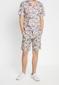 Conjunto de camisa de manga corta y pantalones cortos en un patrón floral. Los colores incluyen rosa, verde y crema. Lleva zapatillas blancas.