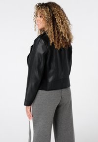 Femme aux cheveux bouclés souriante, portant une veste en cuir noire et un pantalon gris avec une bande blanche, se tenant devant un fond blanc.