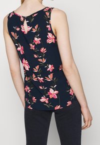 Savisuton sininen toppi, jossa on vaaleanpunaisia kukkakuviota, avokselta takana ja sileä kangas. Ihanteellinen arkikäyttöön.