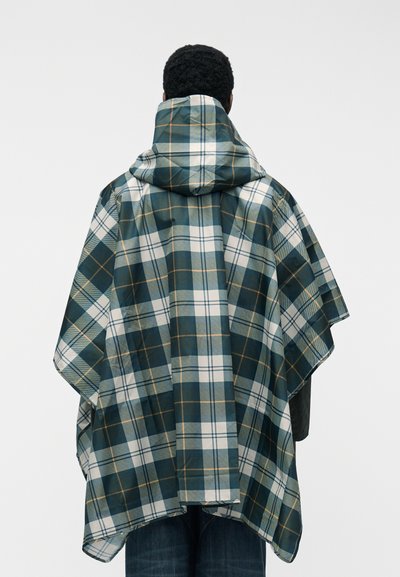 Personne portant un poncho à capuche ivoire, vert et beige à carreaux avec de larges manches, debout contre un fond clair uni, dos à l'appareil photo.