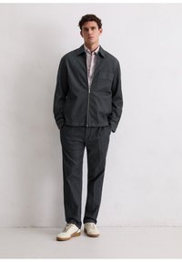 Donkergrijze jas met ritssluiting, puntkraag en borstzak, gecombineerd met bijpassende broek. Draagt witte sneakers. Minimalistisch ontwerp.