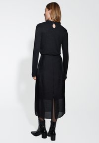 Tenue noire composée d'un haut ajusté avec un dos en trou de serrure et d'une longue jupe fluide avec une fente sur le côté. Fabriquée en tissu léger et texturé.