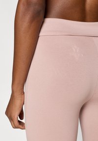 Ροζ κολάν με απαλή υφή και φαρδιά μέση, με διακριτικό λογότυπο και εφαρμοστή σχεδίαση, παρουσιάζοντας μια πλευρική όψη του γοφού.