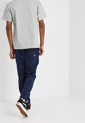 Mand, der går væk iført en lysegrå t-shirt, marineblå bukser med en baglomme og sorte skate-sko med hvide såler.