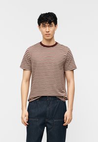 Νέος άνδρας με κοντά σκούρα μαλλιά, φορώντας μια μπλούζα με μπεζ και βυσσινί ρίγες και σκούρο μπλε τζιν, στέκεται μπροστά σε απλό φωτεινό φόντο.