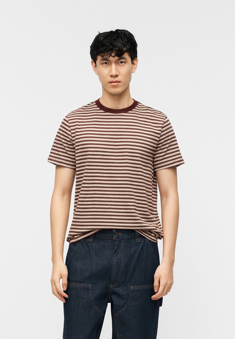 Νέος άνδρας με κοντά σκούρα μαλλιά, φορώντας μια μπλούζα με μπεζ και βυσσινί ρίγες και σκούρο μπλε τζιν, στέκεται μπροστά σε απλό φωτεινό φόντο.