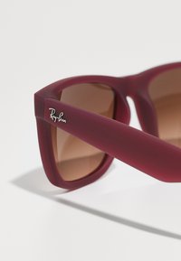 Ochelarii de soare Ray-Ban au un cadru mat de culoare burgundă, lentile maro cu nuanțe gradient și un logo argintiu pe latura templei.