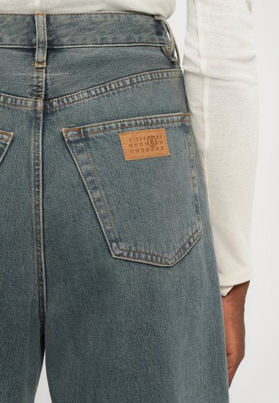 Blue denim jeans with a high waist, a rectangular leather label on the back pocket featuring size numbers, and contrasting orange stitching.