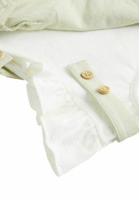Top in cotone verde chiaro sovrapposto a un tessuto bianco sottostante. Presenta bordi arricciati e bottoni in legno su un cinturino decorativo.