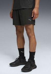 Sorte atletiske shorts med reflekterende Puma-logo, parret med sorte ribbede sokker og sorte løbesko. Teksturen virker let og åndbar.