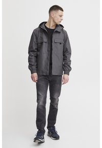 Grå hettejakke med to brystlommer, elastiske mansjetter, kombinert med svart skjorte og falmede grå jeans, fullført med blå joggesko.