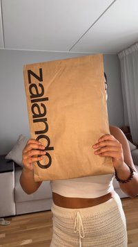 Brązowa koperta z papieru kraft z dużym czarnym logo "Zalando", trzymana przez osobę, lekko pomarszczona tekstura, prostokątny kształt.