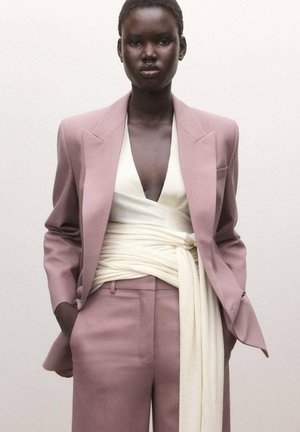 Persona de piel oscura vistiendo un traje lila con una blusa cruzada color crema claro atada a la cintura, de pie con las manos en los bolsillos frente a un fondo liso.