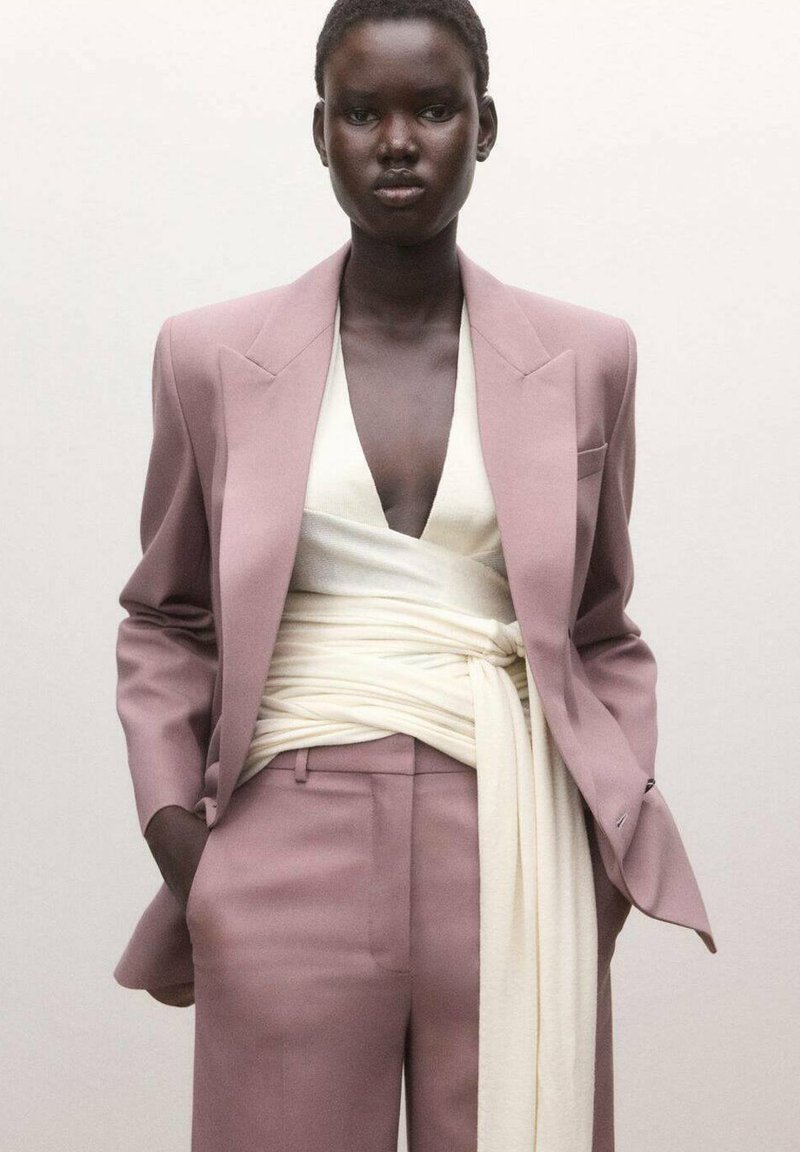 Persona de piel oscura vistiendo un traje lila con una blusa cruzada color crema claro atada a la cintura, de pie con las manos en los bolsillos frente a un fondo liso.