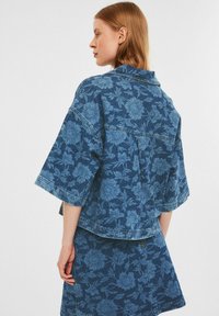 Denimjacke mit floralem Muster in Blau- und Blautönen, mit kurzen Ärmeln, Kragen und lockerer Passform. Der Stoff hat eine strukturierte Oberfläche.
