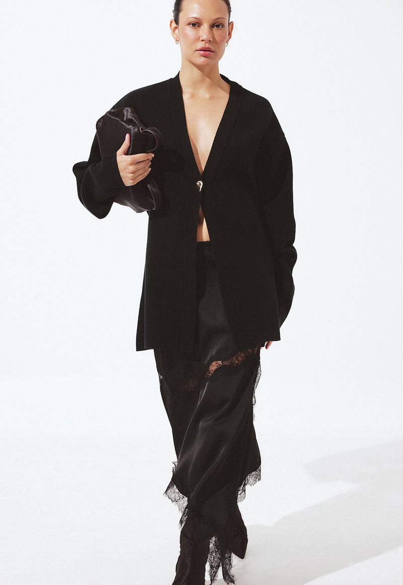 Kobieta w za dużym czarnym kardiganie na czarnej satynowej spódnicy z koronkowym wykończeniem, trzyma czarną kopertówkę, stoi na białym tle.