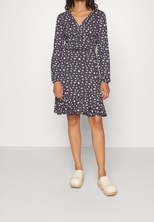 Marineblaue Wickelkleid mit cremefarbenen Pünktchen, langen Ärmeln und Rüschensaum. Kombiniert mit hellen Clogs. Weicher Stoff mit glatter Textur.
