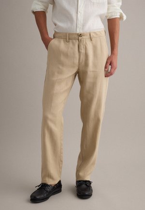 Hombre con pantalones beige de pierna recta, camisa blanca arremangada y zapatos negros de cuero con cordones, de pie con las manos parcialmente en los bolsillos.