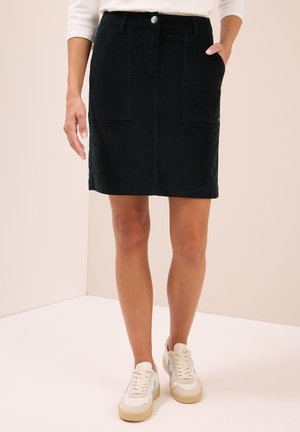 Femme portant une jupe noire à mi-mollet avec de grandes poches avant et des baskets blanches avec des détails bleus, se tenant devant un fond pâle.