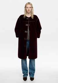 Abrigo de lana burdeos de corte relajado, con frente abotonado y tela texturizada. Combinado con jeans azules y zapatos de punta marrones.