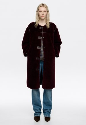 Burgundowa wełniana kurtka o luźnym kroju, z zapięciem na guziki i teksturowanym materiałem. W zestawieniu z niebieskimi dżinsami i brązowymi spiczastymi butami.