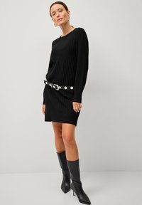 Zwart geribbelde gebreide jurk met lange mouwen, strakker rond de taille door een contrasterende riem met grote zilveren studs. Zwarte laarzen met hak tot aan de knie.