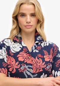 Mujer rubia con piel clara que lleva una camisa de manga corta azul marino con patrones florales tropicales en rojo y blanco, con los brazos cruzados frente a ella.