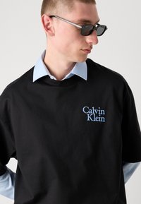 T-shirt en coton noir présentant un logo brodé "Calvin Klein" en bleu clair. En dessous, une chemise à col bleu clair est partiellement visible.