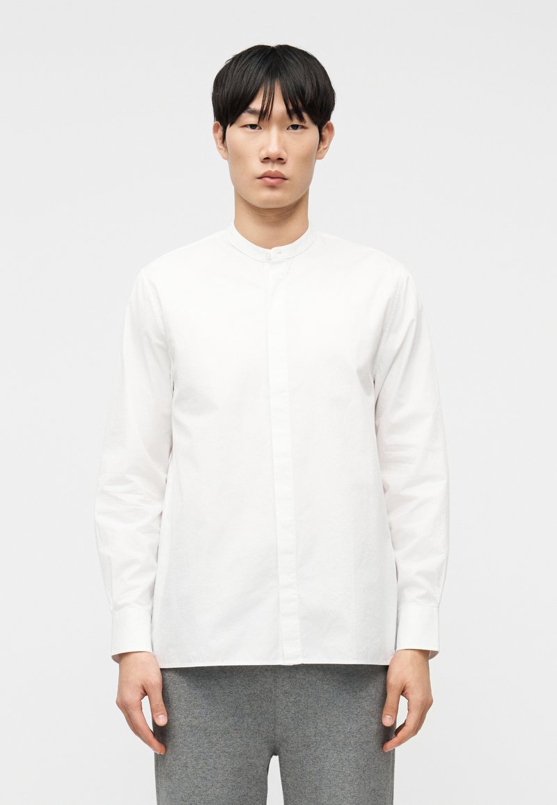 Νεαρός άνδρας που φοράει λευκό που κάμισο με μανδρινή γιακά και γκρι παντελόνι, στέκεται μπροστά σε απλό ανοιχτό φόντο.