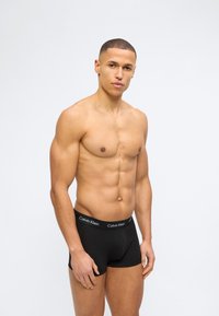 Ευκίνητος νέος άνδρας όρθιος, φορώντας μαύρο μποξεράκι Calvin Klein, με ουδέτερη έκφραση μπροστά σε απλό λευκό φόντο.