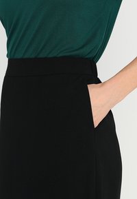 Gonna nera con una texture liscia e tasche laterali. Abbinata a una maglia verde scuro, che illustra una silhouette aderente senza decorazioni.