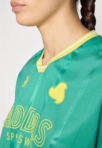 Green sports jersey featuring a yellow V-neck; accented with a yellow logo and stripe details. Lightweight, textured fabric.