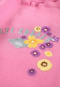 Tissu rose avec le texte argenté scintillant "LEE COOPER" et des motifs floraux colorés en jaune, violet et blanc. Présente un tissu texturé.