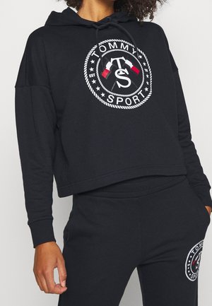 Osoba nosząca czarną krótka bluzę z kapturem i spodnie z białym i czerwonym logo Tommy Sport przedstawiającym skrzyżowane flagi i motyw koła z liny.