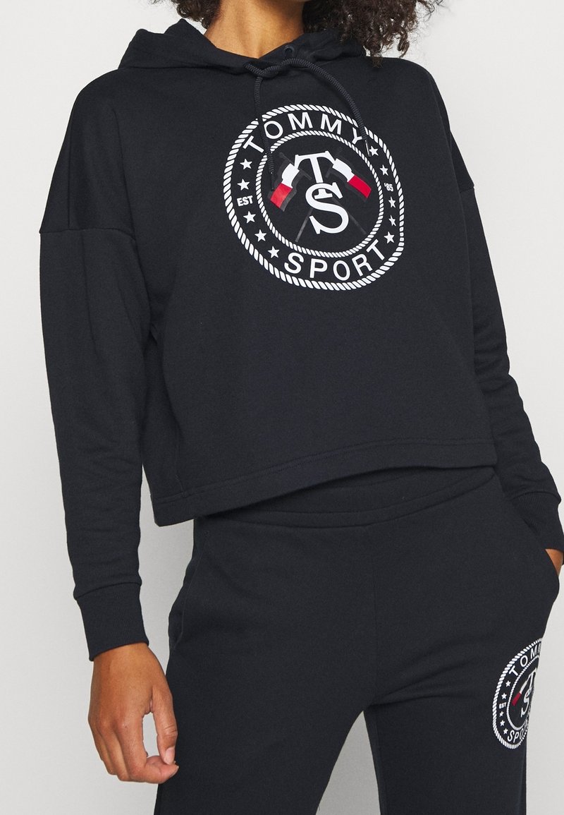 Personne portant un sweat à capuche court noir et un pantalon avec le logo Tommy Sport blanc et rouge présentant des drapeaux croisés et un motif de cercle en corde.