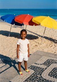 Junges Kind in weißem Outfit und bunten Sandalen steht auf gemustertem Pflaster neben einem Sandstrand mit blauen, roten und gelben Sonnenschirmen.