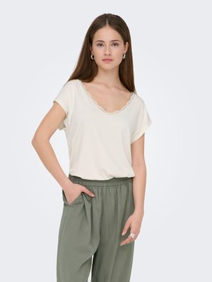 Krämfärgad T-shirt med en V-ringning prydd med spetskant, kombinerad med löst sittande olivgröna byxor med elastisk midja och sidofickor.