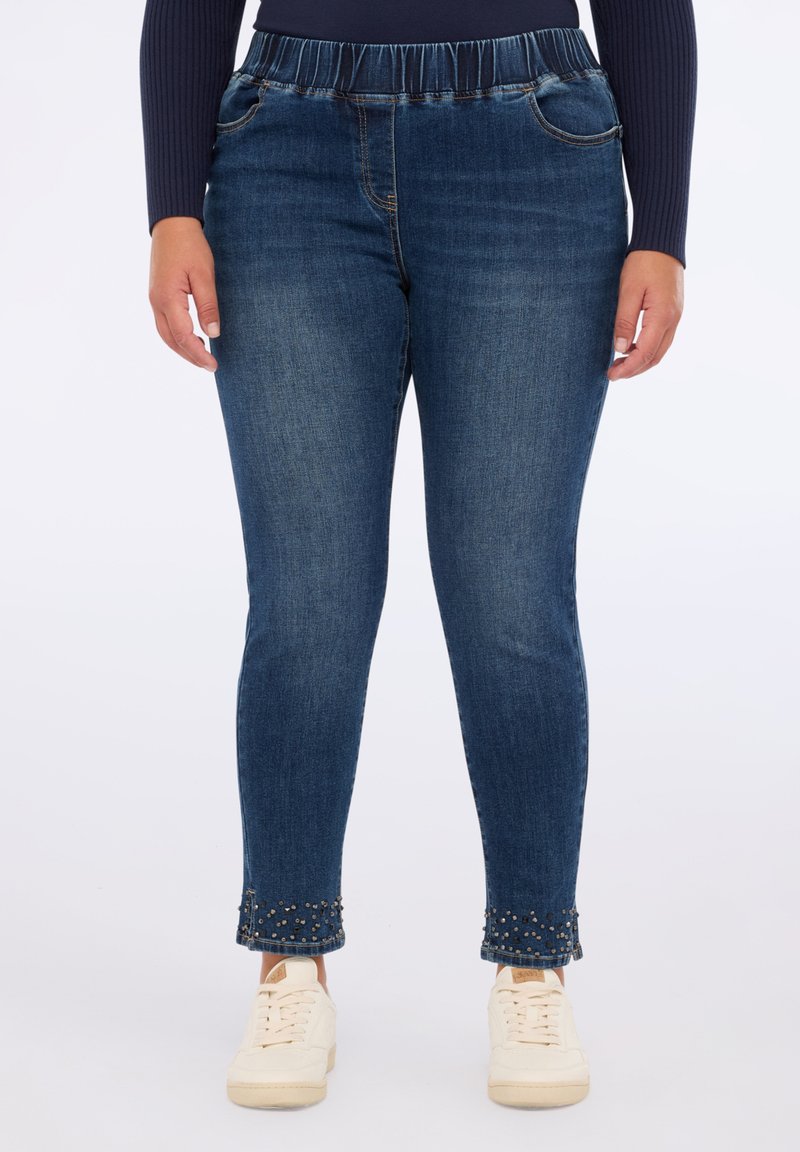 Jeans in denim con cintura elastica arricciata, fit aderente e orli decorati con borchie metalliche. Lavaggio blu medio, texture liscia.