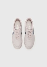Adidași Nike roz deschis cu o textură netedă, swoosh gri, șireturi plate și tălpi cu amortizare, având un design minimalist.