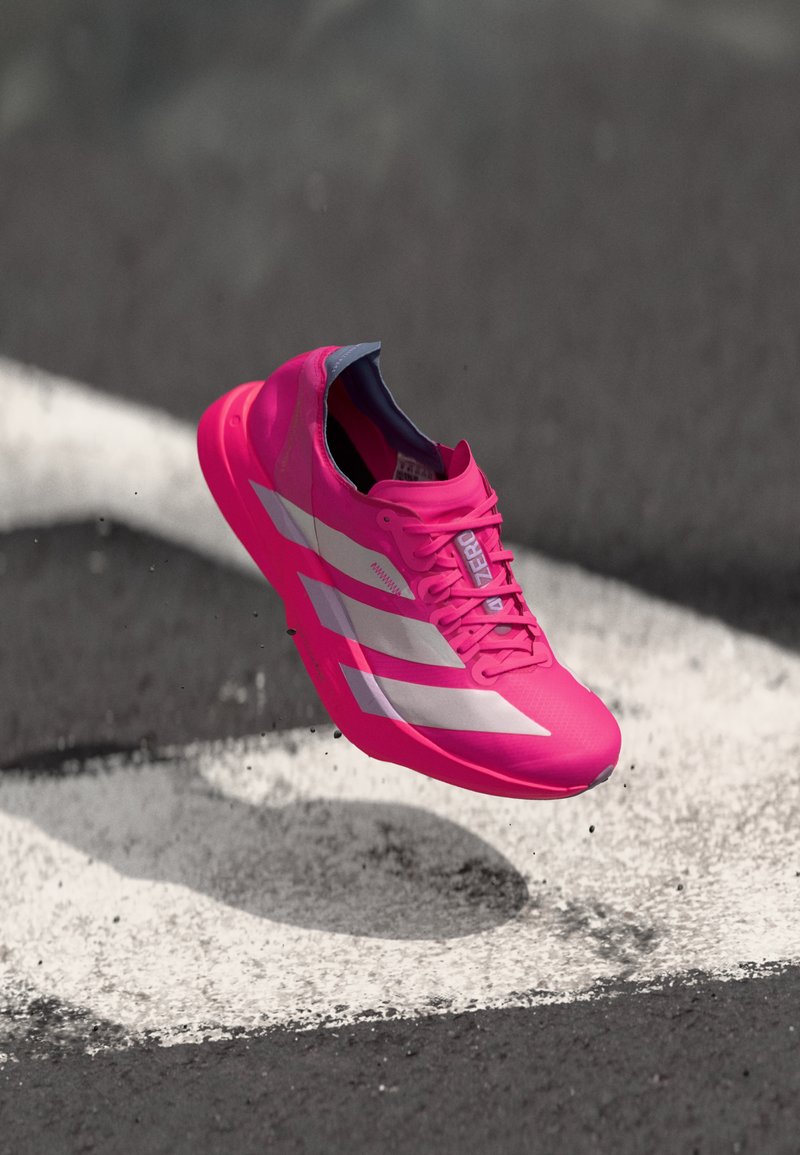 Sapatilha de desporto de cor rosa-brilhante com um design elegante, apresentando riscas brancas e uma superfície texturizada, pairando acima de uma linha branca no asfalto.