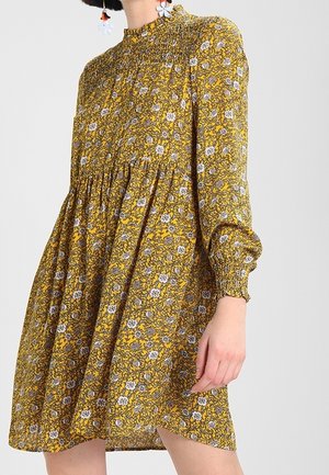 Femme portant une robe longue jaune à fleurs avec un col montant froncé et des poignets, debout devant un fond clair uni.
