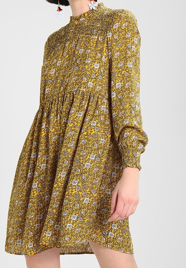 Femme portant une robe longue jaune à fleurs avec un col montant froncé et des poignets, debout devant un fond clair uni.